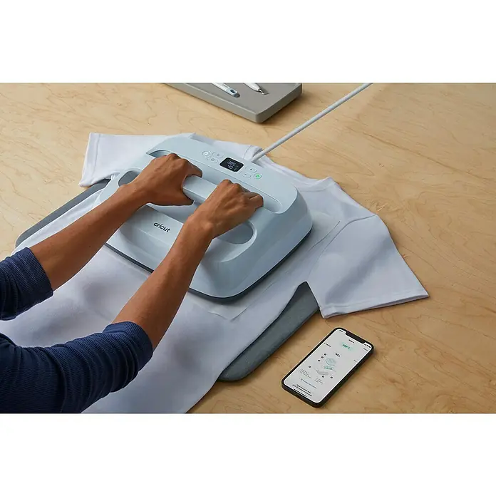 Cricut Transferpresse Easypress 3 - B x L: 30 x 25 cm, Passend für: Bügelfolien, Drucktechnologie: Wärmeübertragung - Eine Person benutzt eine Cricut EasyPress 2 auf einem weißen T-Shirt, mit einem Smartphone daneben.