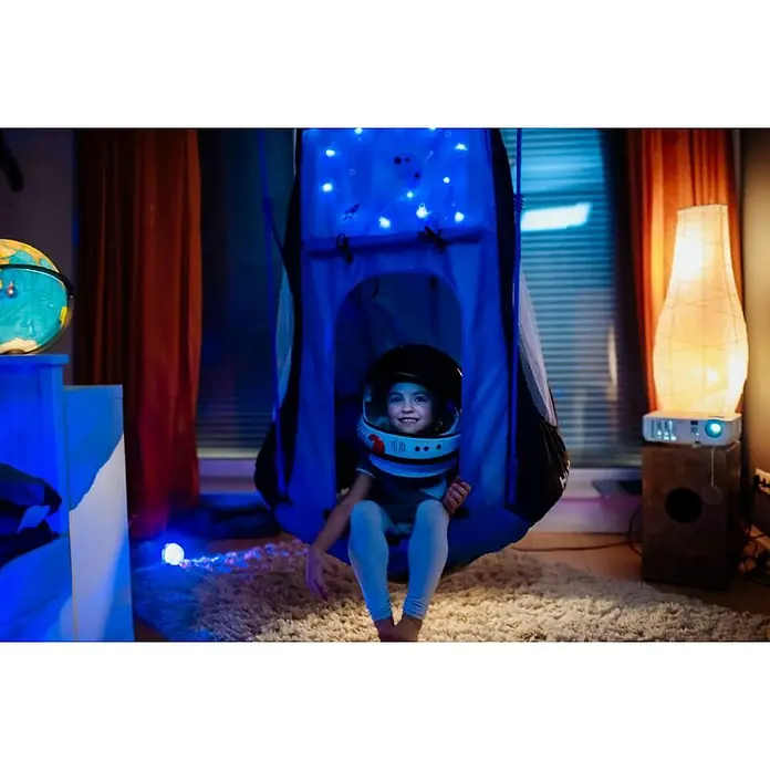 Hudora Nestschaukel Cosmos LED 90 - Durchmesser: 90 cm, Glas, Blau, Schwarz - Ein Kind im Astronautenhelm sitzt in einer Hängematte, die wie ein Raumschiff aussieht, umgeben von Sternenlichtern.