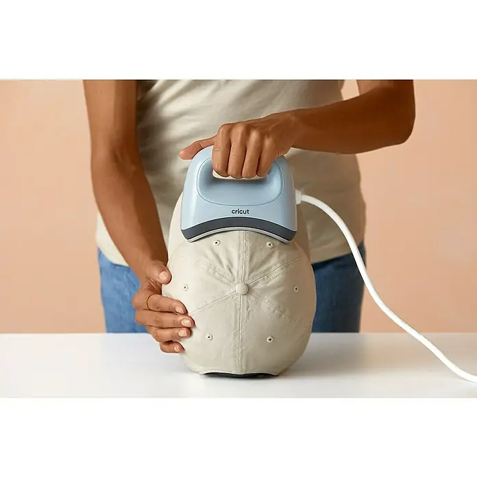 Cricut Transferpresse Hat Press - Passend für: Mützen, Drucktechnologie: Wärmeübertragung - Nahaufnahme einer Person, die mit einer Cricut-Hitzepresse ein Design auf eine beigefarbene Baseballkappe aufbringt, ideal für DIY-Projekte.