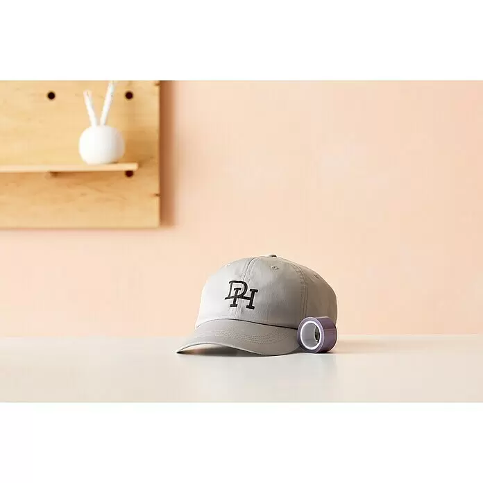 Cricut Klebeband Strong Heat Resistant - 10 m x 2,5 cm - Graue Baseballkappe mit schwarzem „PI“-Logo und einer Rolle lila Klebeband auf heller Fläche, im Hintergrund eine verschwommene Holzlochwand.