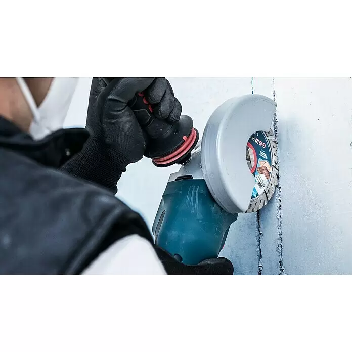 Bosch Professional X-Lock Diamant-Trennscheibe Expert for MultiMaterial - Durchmesser Scheibe: 125 mm - Nahaufnahme einer Person in schwarzen Handschuhen, die einen blaugrauen Winkelschleifer mit Trennscheibe an einer hellen Wand bedient.