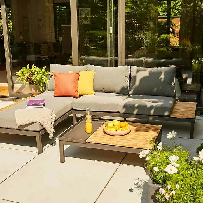Siena Garden Loungemöbel-Set Alvida - 3 -tlg., Anthrazit/Schwarz, Teakholz, Tablett, Max. Personenzahl: 3 - Eine stilvolle graue Gartenlounge mit farbenfrohen Kissen und einem passenden Tisch mit Obst und Getränken, ideal zum Entspannen im Freien.