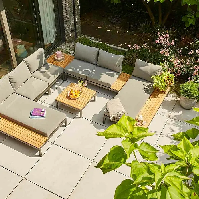 Siena Garden Loungemöbel-Set Alvida - 5 -tlg., Anthrazit/Schwarz, Teakholz, Tablett, Max. Personenzahl: 4 - Eine moderne Gartensitzgruppe mit grauen Polstern und Holzelementen steht auf einer Terrasse, umgeben von üppigem Grün und Blumen.