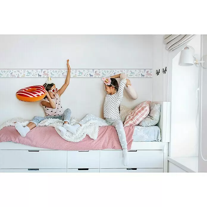 Marburg Kids' Walls Tapetenborte Einhörner - Weiß/Multicolor, Motiv, L x H: 5 x 0,18 m, Vlies - Zwei Mädchen spielen auf einem weißen Bett mit integrierten Schubladen, unter einer Wand mit bunter Einhorn-Tapetenbordüre.