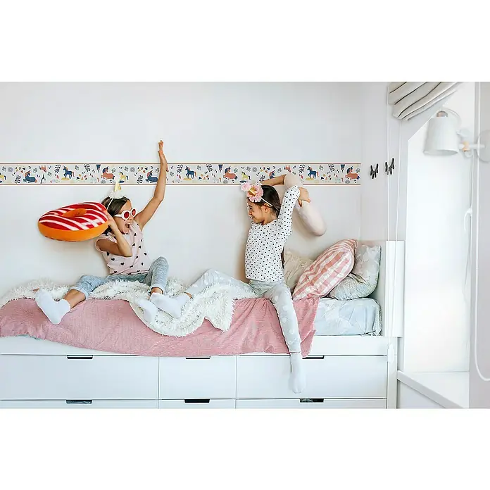 Marburg Kids' Walls Tapetenborte Einhörner - Grau/Multicolor, Motiv, L x H: 5 x 0,18 m, Vlies - Zwei Mädchen in einem Kinderzimmer spielen auf einem Bett mit weißem Bettkasten und einer Bordüre mit Einhorn-Motiv an der Wand.