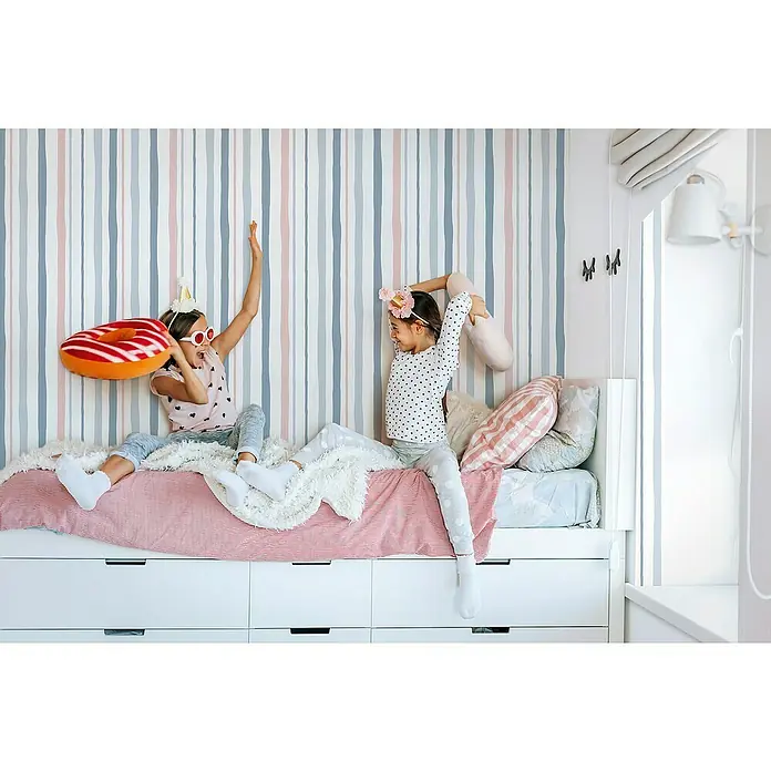 Marburg Kids' Walls Vliestapete Kombistreifen II - Weiß/Grau/Rosa, Streifen, 10,05 x 0,53 m - Zwei spielende Mädchen auf einem Bett mit Schubladen, das mit einer rot gestreiften Decke und Kissen dekoriert ist.