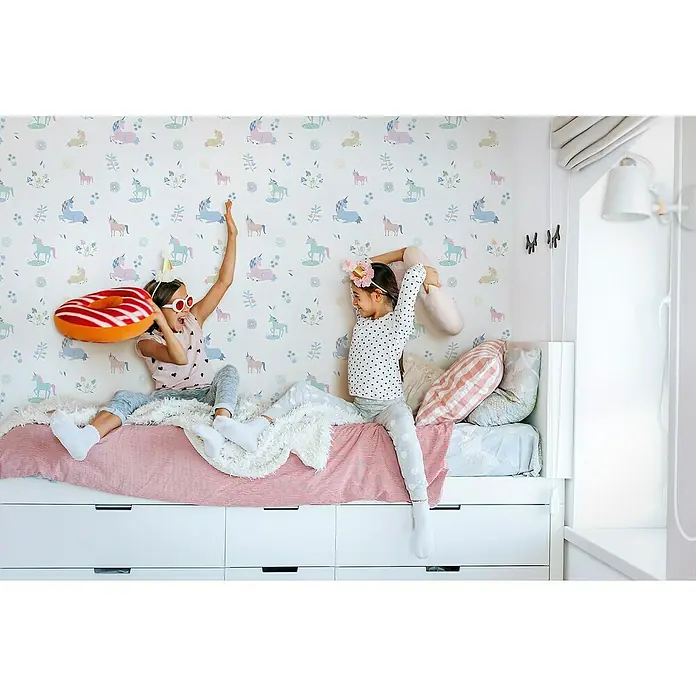 Marburg Kids' Walls Vliestapete Einhörner - Weiß/Multicolor, Tieroptik, 10,05 x 0,53 m - Zwei Mädchen spielen auf einem Bett mit Schubladen, dekoriert mit einem bunten Einhornmuster. Ein Spaß für Kinder!