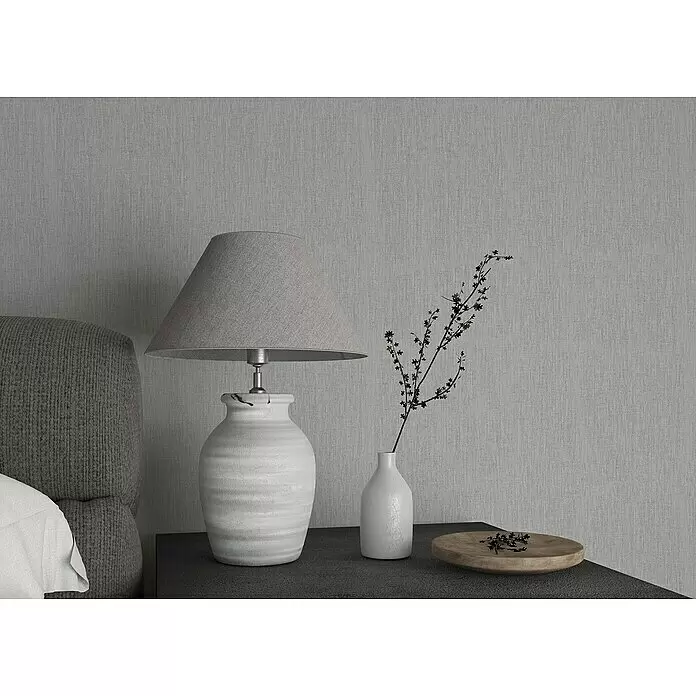 Marburg Botanica Vliestapete - Grau, Uni, 10,05 x 0,53 m - Eine gemütliche Nachttischszene mit einer grauen Lampe, einer weißen Vase mit Zierzweigen und einer Holzschale.