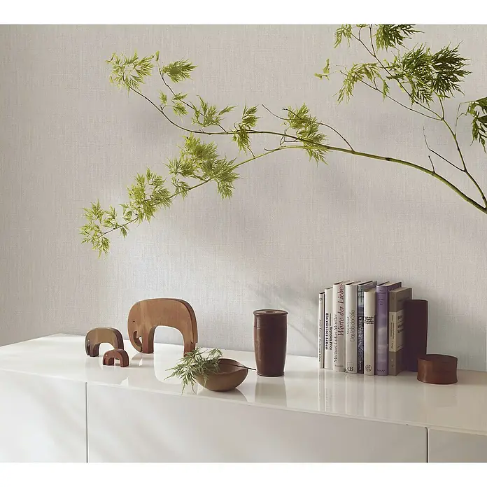 Marburg Botanica Vliestapete Struktur - Greige, Uni, 10,05 x 0,53 m - Dekorative Anordnung mit Büchern, Holzelefanten, einer Pflanze und einem Behälter auf einer weißen Kommode.