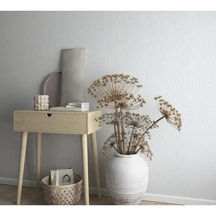 Marburg Botanica Vliestapete Struktur - Greige, Uni, 10,05 x 0,53 m - Eine elegante Komposition mit einem hellen Holzbeistelltisch, einer weißen Vase mit getrockneten Blumen und dekorativen Objekten vor einer strukturierten Wand.