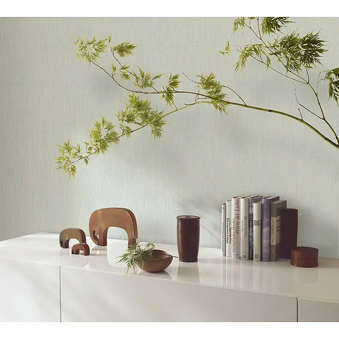 Marburg Botanica Vliestapete Struktur - Graugrün, Uni, 10,05 x 0,53 m - Nahaufnahme eines dekorierten, weißen Sideboards mit Holzelefanten, Bücher und einer grünen Pflanze vor einer hellen Wand.
