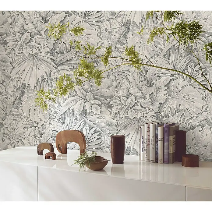 Marburg Botanica Vliestapete - Hellgrau, Floral, 10,05 x 0,53 m - Nahaufnahme eines weißen Sideboards mit Dekorationen wie Holzelefanten, Bücher und Grünpflanzen vor einer floralen Tapete.