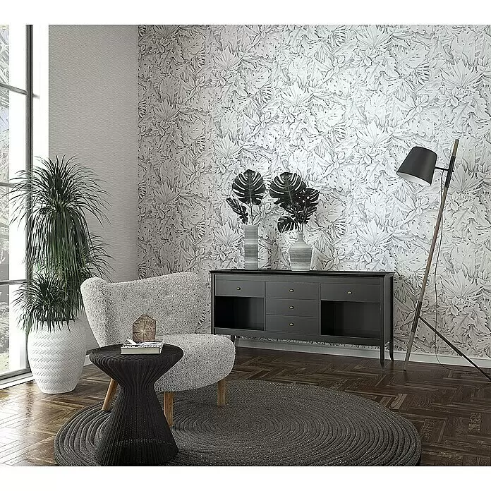 Marburg Botanica Vliestapete - Hellgrau, Floral, 10,05 x 0,53 m - Ein modernes Wohnzimmer mit grauer Blattmuster-Tapete, einem gemütlichen Sessel, einem runden geflochtenen Teppich und einem schwarzen Konsolentisch mit Pflanzendekor.