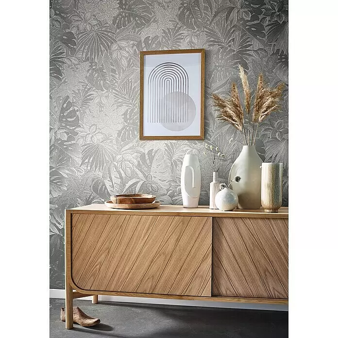Marburg Botanica Vliestapete - Silber, Floral, 10,05 x 0,53 m - Helles Holz-Sideboard mit schrägen Schiebetüren, dekoriert mit Vasen und Schalen, in einem Raum mit gemusterter Tapete.