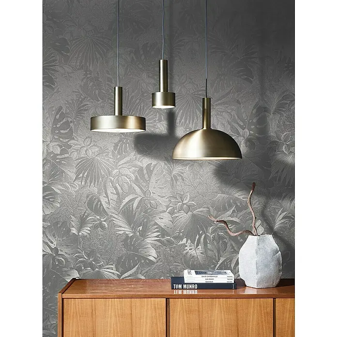 Marburg Botanica Vliestapete - Silber, Floral, 10,05 x 0,53 m - Drei moderne Pendelleuchten aus gebürstetem Messing, die über einem Holzschrank mit Dekorationen hängen, vor einer grauen Blattmustertapete.
