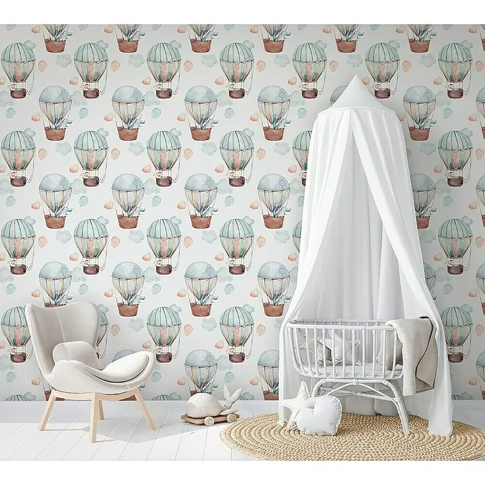 Marburg Kids' Walls Vliestapete Heißluftballons - Weiß/Blau/Beige, Grafisch, 10,05 x 0,53 m - Kinderzimmer mit Akzentwand mit Heißluftballons, ein cremefarbener Sessel und eine weiße Babywiege mit Überdachung.