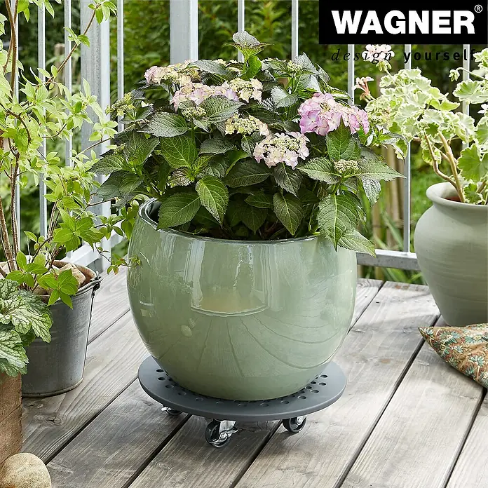 Wagner System Pflanzenroller - 34 cm, Beton, Anthrazit, Max. Belastung: 100 kg - Ein runder, grüner Pflanztopf mit rosafarbenen Blüten steht auf einem Rollbrett auf einer Holzterrasse.