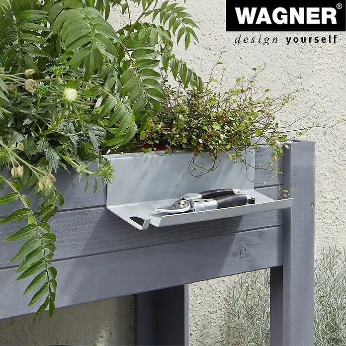 Wagner System GreenFamily Ablagekonsole GH 3001 - Passend für: Wagener GreenFamily Hochbeete, Stahl - Nahaufnahme eines grauen Pflanzkastens mit Schere auf einer Ablagefläche, umgeben von üppigem Grün und dem Wagner-Logo.