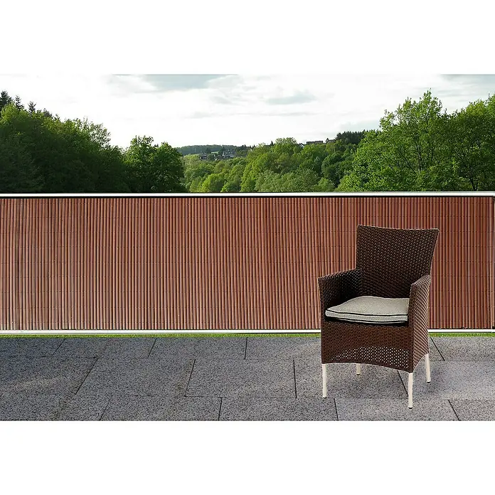 Gardol Comfort Sichtschutzmatte - Kastanienbraun, 300 x 90 cm - Brauner Rattansessel mit Sitzkissen auf einer Terrasse mit Blick auf grüne Bäume im Hintergrund. Das Objekt ist auf einem grauen Steinboden positioniert.