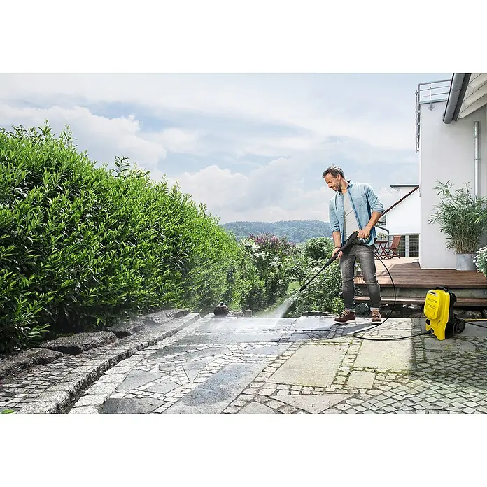 Kärcher Hidrolimpiadora de alta presión K 4 Compact UM - 130 bar, 1.800 W, Capacidad de bombeo: 420 l/h - Un hombre limpia un patio con una hidrolavadora Kärcher amarilla, eliminando la suciedad.