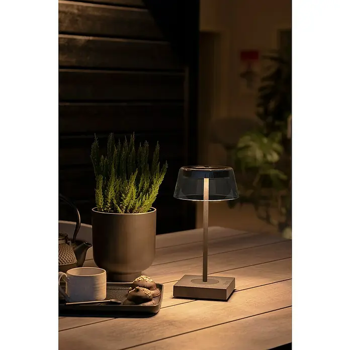 Konstsmide Akku-Tischleuchte Scilla - 2,5 W, L x B x H: 11 x 11 x 27 cm, Schwarz, Warmweiß - Tischdekoration im Freien mit einer eleganten Lampe, die warmes Licht spendet. Eine Tasse, Gebäck und eine Pflanze ergänzen die Szene.