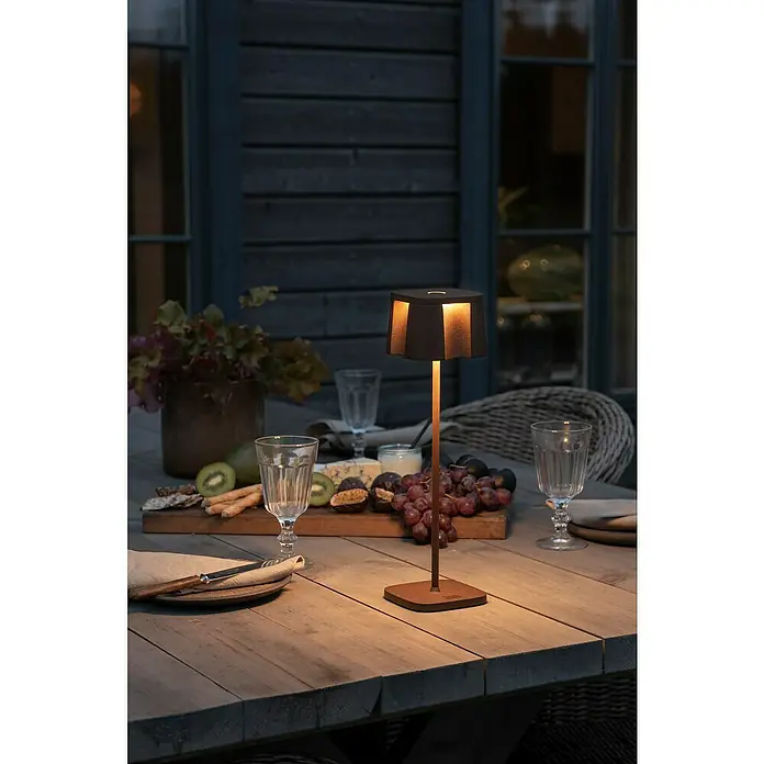 Konstsmide Akku-Tischleuchte Nice - 2,5 W, L x B x H: 10 x 10 x 36 cm, Rost, Warmweiß - Eine Tischlampe aus dunklem Metall mit warmem Licht, die auf einem Tisch mit Speisen und Getränken steht, sorgt für eine gemütliche Atmosphäre.