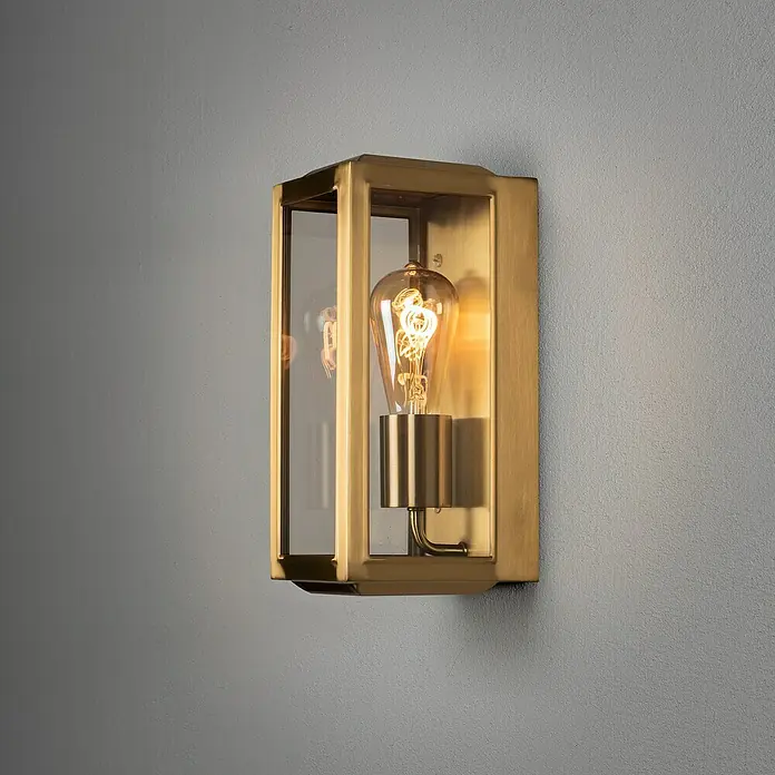 Konstsmide Außenwandleuchte Carpi - 60 W, 15 x 12,5 x 30 cm, Messing, IP44 - Nahaufnahme einer rechteckigen goldenen Wandleuchte mit Glaseinsatz und einer Edison-Glühbirne an einer grauen Wand.