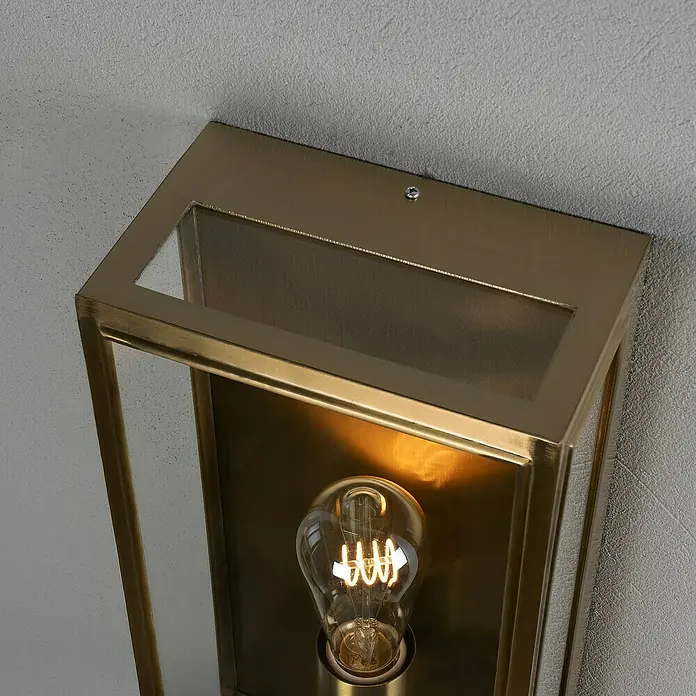 Konstsmide Außenwandleuchte Carpi - 60 W, 12,5 x 20,5 x 40 cm, Messing, IP44 - Nahaufnahme einer modernen Wandleuchte mit goldfarbenem Metallrahmen und einer Edison-Glühbirne, die warmes Licht ausstrahlt.