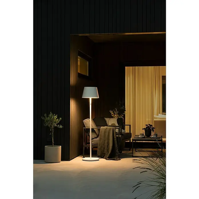 Konstsmide Außenleuchte Pomezia - Akkubetrieben, 10 W, RGBW, 35 x 35 x 150 cm, Mit Leuchtmittel, Weiß - Eine elegante Stehlampe beleuchtet eine stilvolle Terrasse mit Sitzgelegenheiten und Pflanzen. Perfekt für entspannte Abende im Freien.