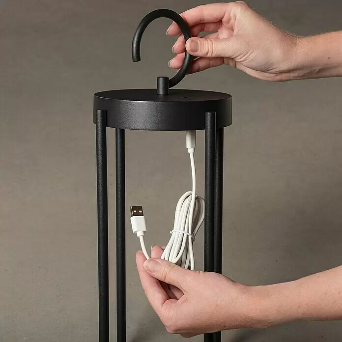 Konstsmide Außenlaterne Manorola - 15,5 x 15 x 39 cm, Schwarz - Hände befestigen einen gebogenen Griff an einer schwarzen Handleuchte und halten das weiße USB-Ladekabel.