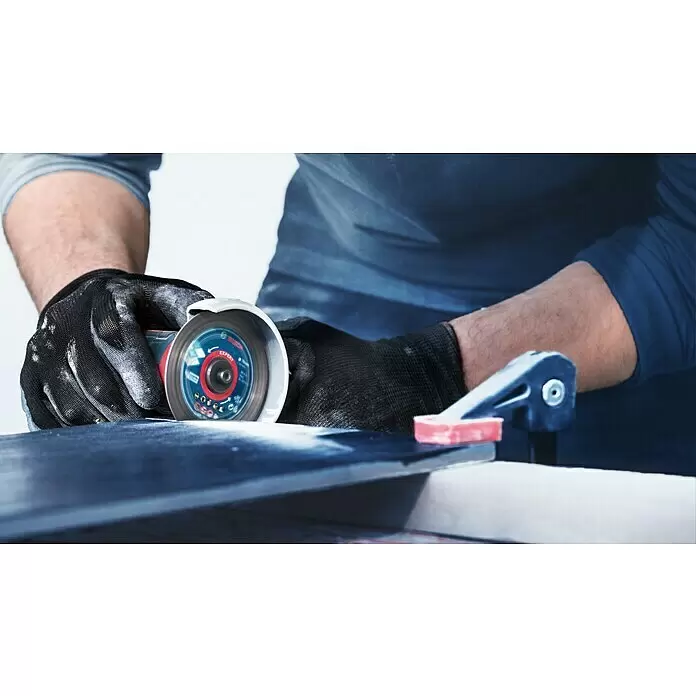 Bosch Expert Diamant-Trennscheibe Hard Ceramic - Geeignet für: Fliesen, Durchmesser Scheibe: 76 mm, 1 Stk. - Hände in schwarzen Handschuhen verwenden einen Winkelschleifer mit Bosch-Scheibe an einer dunklen Platte, gesichert mit einer blauen Klemme.