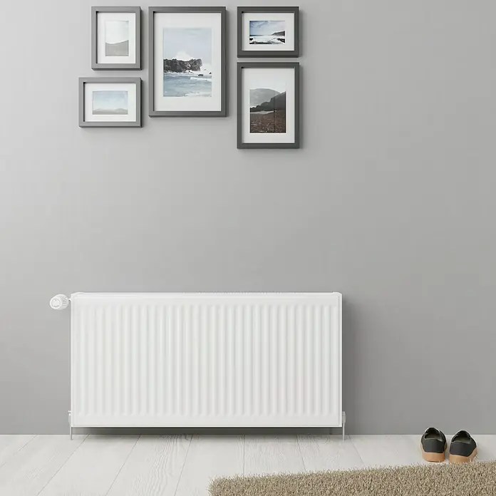Kompaktheizkörper - B x H: 1.000 x 900 mm, Wärmeleistung bei Vorlauftemperatur von 75 °C (D: 50): 2.470 W, Typ Heizkörper: 22 - Nahaufnahme eines weißen Heizkörpers mit Thermostat und sechs gerahmten Bildern an einer grauen Wand.