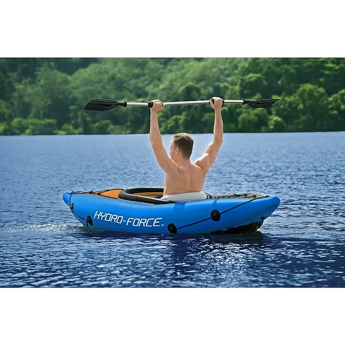 Bestway Hydro Force Kayak Cove Champion - Plástico, Azul/Naranja/Blanco - Un hombre rema en un kayak azul Hydro-Force en un lago, con los brazos levantados y la pala arriba. El kayak se desliza suavemente por el agua.