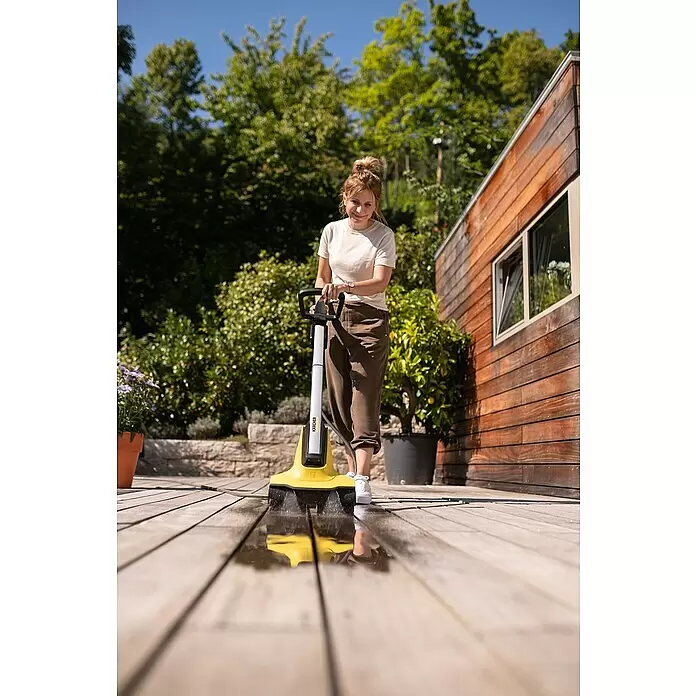 Kärcher Flächenreiniger PCL 3-18 - 18 V, 2,5 Ah, Ohne Akku, 10 bar - Gelb-weißer Kärcher Terrassenreiniger wird von einer Frau verwendet, besprüht eine Holzterrasse mit Wasser.