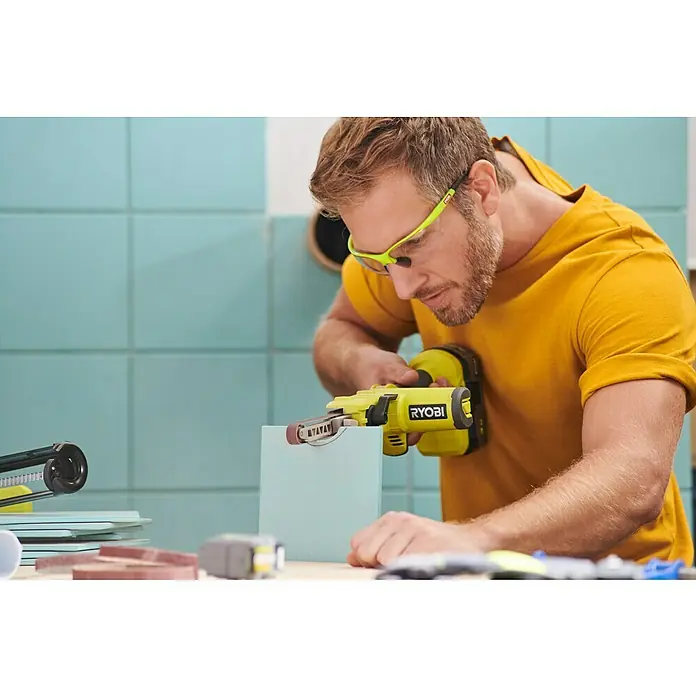 Ryobi ONE+ Maschinenpaket - Akku-Bandschleifer R18PF-0 + gratis Starter-Kit - Ein Mann verwendet einen gelben Ryobi-Bandschleifer, um ein Werkstück zu bearbeiten. Er trägt eine gelbes T-Shirt und eine Schutzbrille.