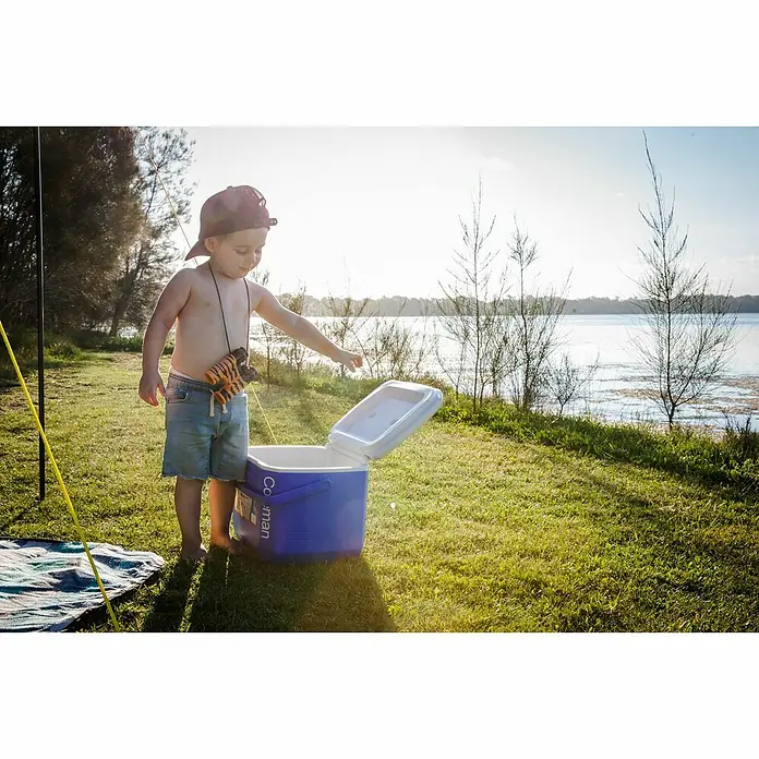 Coleman Nevera de camping 16QT - L x An x Al: 31 x 39 x 27 cm, Azul, 15 l, Asa de transporte ergonómica - Un niño pequeño está de pie junto a una nevera portátil azul Coleman al aire libre, listo para un picnic o una aventura.