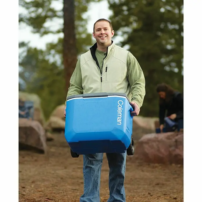 Coleman Nevera de camping 60QT - L x An x Al: 44 x 57 x 45 cm, Azul, 56 l, Ruedas - Un hombre lleva una nevera azul Coleman con ruedas al aire libre. Ideal para picnics y camping.