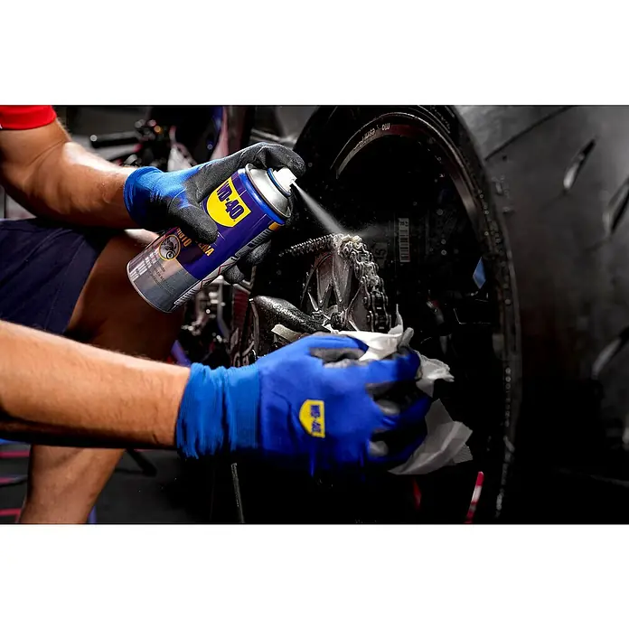 WD-40 Specialist Motorbike Grasa para cadena - 400 ml - Una persona rociando limpiador de cadenas de moto WD-40 en la cadena de una moto, mientras la limpia con un paño.