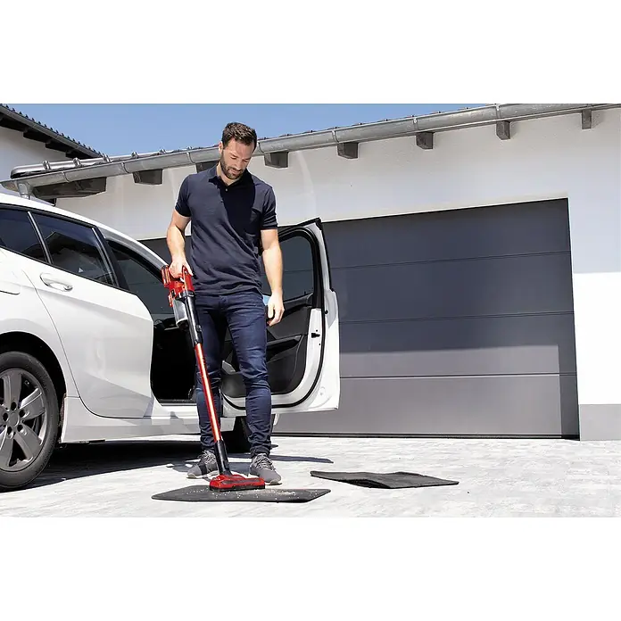 Einhell Power X-Change 18V Akku-Staubsauger TE-SV 18 Li-Solo - 18 V, Ohne Akku, 4 Ah, Behältervolumen: 0,6 l - Mann saugt mit einem roten Akku-Staubsauger eine Fußmatte vor einem weißen Auto und einer Garage.