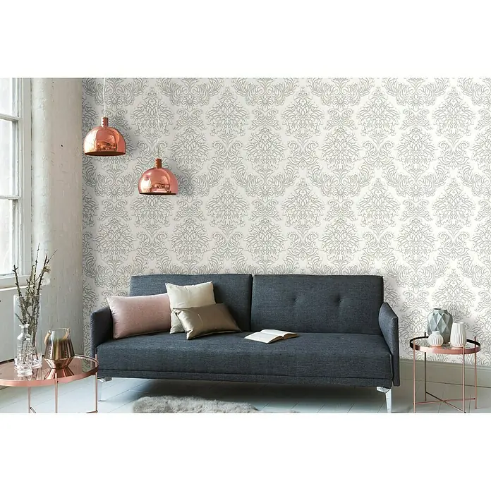 A.S. Création Metropolitan Stories Vliestapete DamaskAltweiß, Ornament, 10,05 x 0,53 m - Ein graues Sofa mit Kissen und Decken steht vor einer Wand mit einem eleganten grauen Damastmuster. Die Kupferleuchten verleihen dem Raum einen Hauch von Wärme.