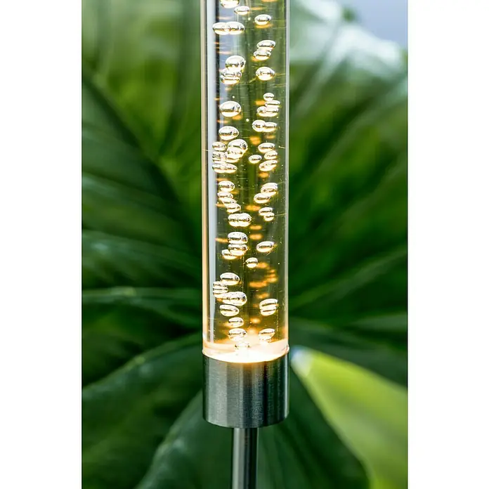 BAUHAUS Lámpara decorativa solar Bubble - 0,06 W, Transparente, IP44 - Primer plano de una luz solar de jardín en forma de tubo con burbujas de aire y una base de metal sobre un fondo verde.