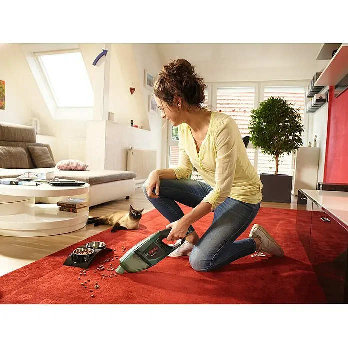 Bosch Power for All 12V Akku-Handstaubsauger EasyVac 12 - 12 V, Ohne Akku - Eine Frau kniet auf einem roten Teppich und saugt mit einem Bosch-Akkusauger Futterreste auf, während eine Katze zusieht.