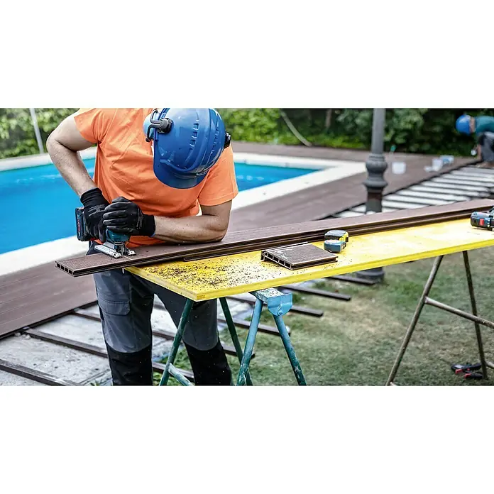 Bosch Stichsägeblätter Plastic Composites Clean T 301 CHM - T-Schaft - Ein Handwerker sägt mit einer Bosch Stichsäge ein braunes Terrassendiele auf einem gelben Tisch. Er trägt Arbeitskleidung und einen blauen Schutzhelm.