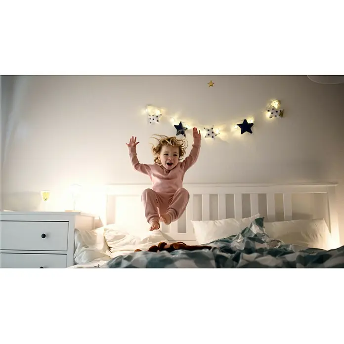Garza Set de bombillas LED Filamento - E27, Blanco cálido, 1.050 lm, 8 W, 3 ud. - Una niña alegre salta en una cama decorada con una guirnalda de estrellas, almohadas suaves y un edredón acogedor, creando un ambiente lúdico.