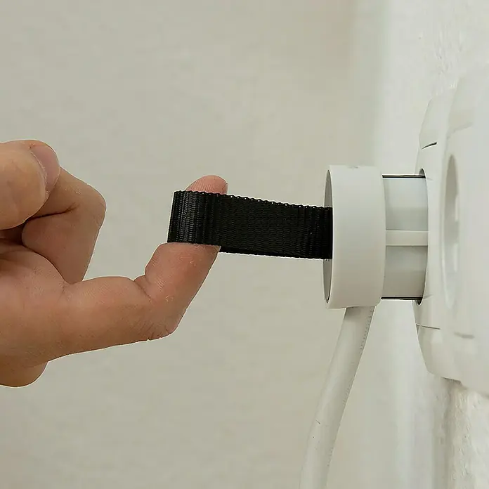 Simon Base aérea móvil - IP20, Blanco - Primer plano de una mano que tira de un enchufe con una cinta negra de una toma de corriente blanca, diseñado para fácil extracción.