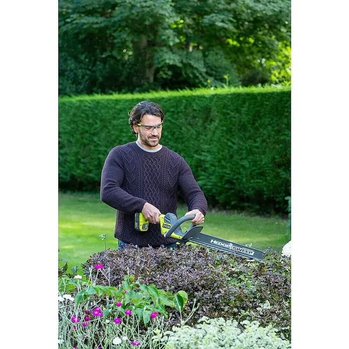 Ryobi ONE+ Akku-Heckenschere RY18HT45A-0 - 18 V, Li-Ionen, Ohne Akku - Ein Mann benutzt eine RYOBI Akku-Heckenschere, um Büsche im Garten zu schneiden, wobei er eine Schutzbrille trägt.