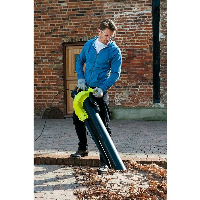Ryobi Elektro-Laubbläser RBV3000CSV - 3.000 W, Blasgeschwindigkeit: 375 km/h, Blasleistung: 14 m³/min - Ein Mann benutzt einen Laubbläser, um Blätter von einem Backsteinweg zu entfernen. Der gelbe Laubbläser von Ryobi wird mit Strom betrieben.