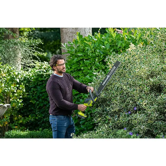 Ryobi ONE+ Cortasetos de batería RY18HT50A-0 - 18 V, Iones de litio, Sin batería - Un hombre usa un cortasetos Ryobi para podar un seto verde, usando gafas de seguridad para proteger sus ojos.