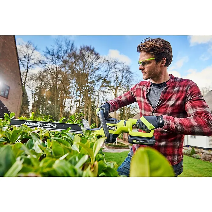 Ryobi Max Power Akku-Heckenschere RY36HTX65A-0 - 36 V, Li-Ionen, Ohne Akku - Ein Mann mit Schutzbrille benutzt eine Ryobi Akku-Heckenschere, um eine Hecke zu schneiden, mit Bäumen und Häusern im Hintergrund.