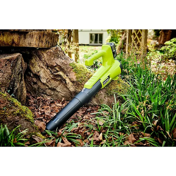 Ryobi ONE+ Soplador de hojas de batería RY18BLA - 18 V, Sin batería, Velocidad de soplado: 145 km/h, Potencia de soplado: 7,1 m³/min - Un soplador de hojas inalámbrico Ryobi verde y negro descansa junto a un tocón de árbol, rodeado de hojas y vegetación.